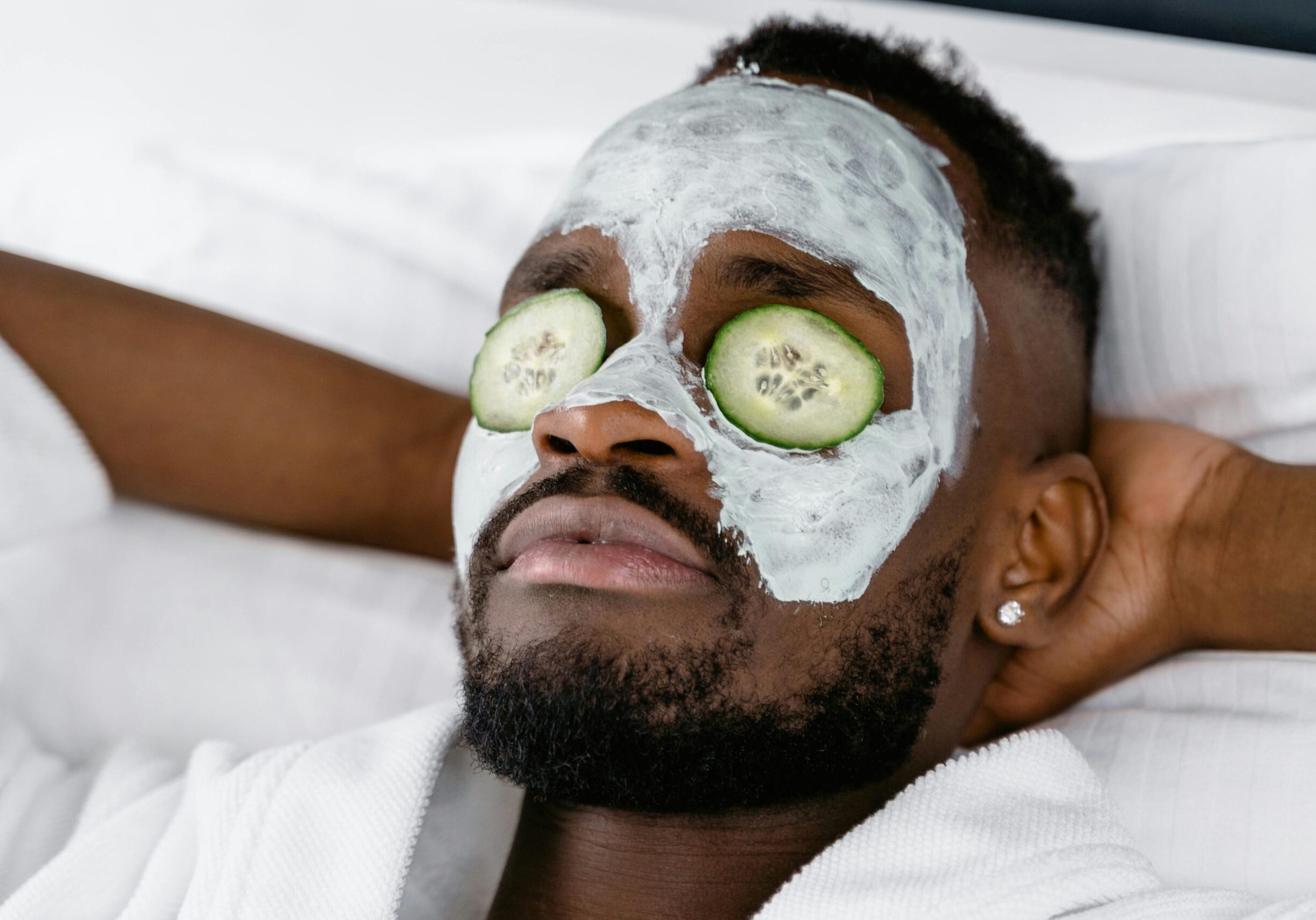Image of a man reclining with his hands behind his head, facial mask on his face and cucumbers over his eyes.